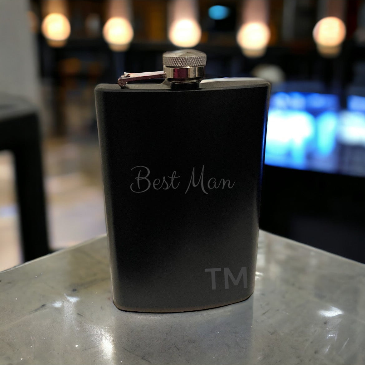 A stainless steel hip flask with 'Best Man' text and a small symbol engraved on the front, placed on a reflective surface.