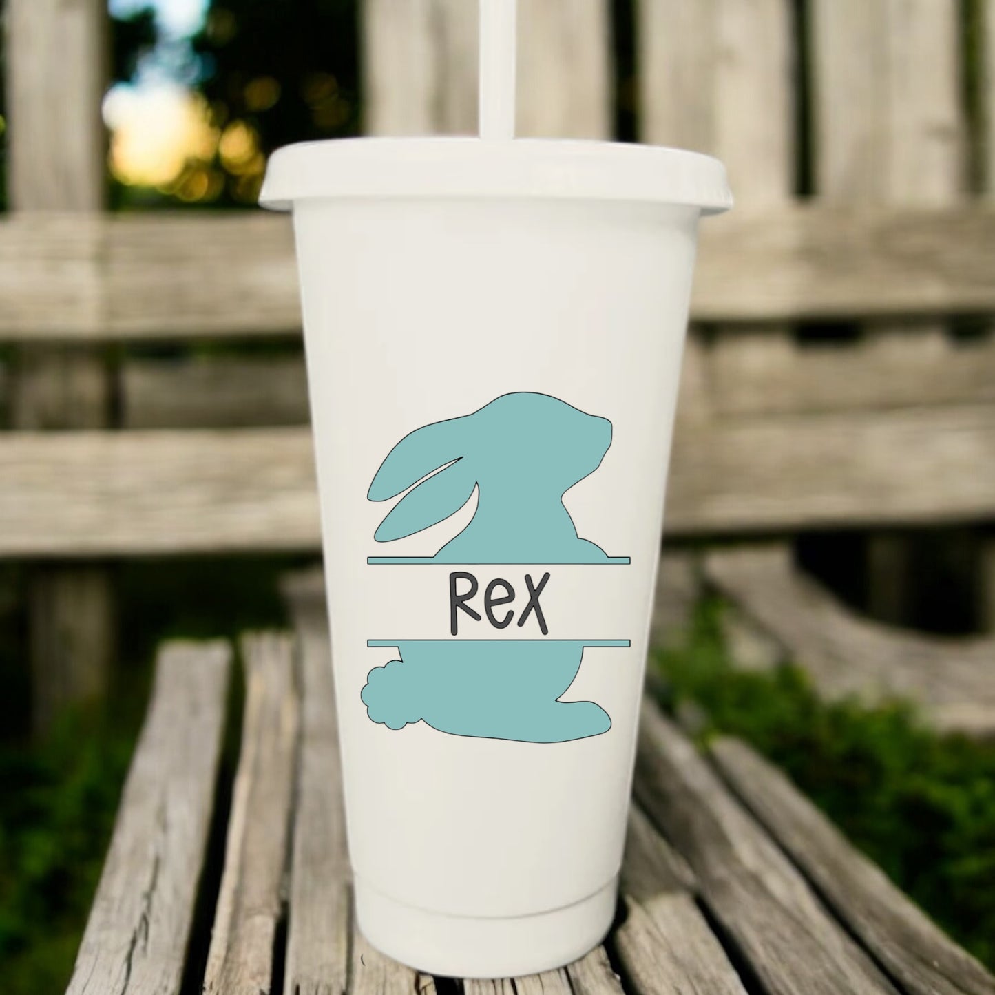 A 24oz white cup with a mint green bunny and the name 'Rex' printed on it, placed on a wooden bench with a drawstring sack.