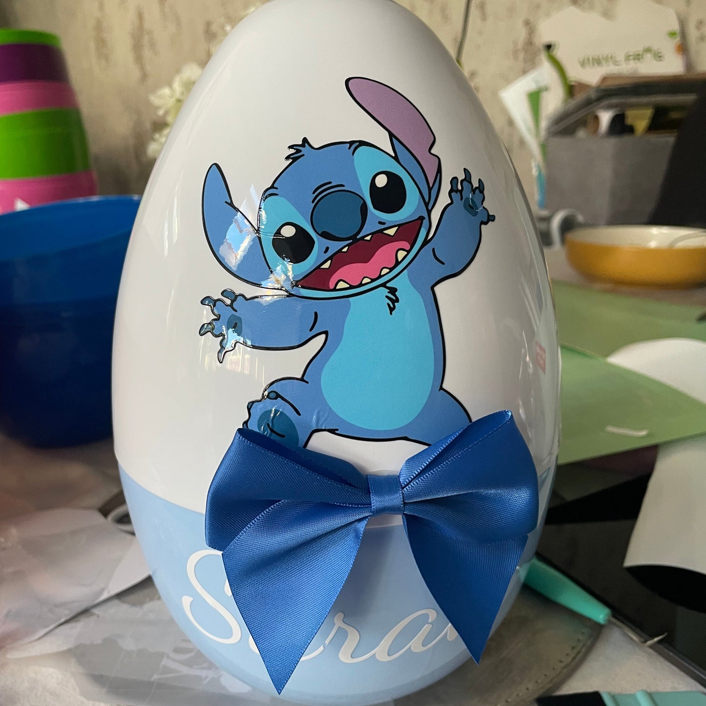 A large, white, fillable Easter egg with a cartoon character resembling Stitch from Lilo and Stitch depicted on it, with a blue bow tie added.