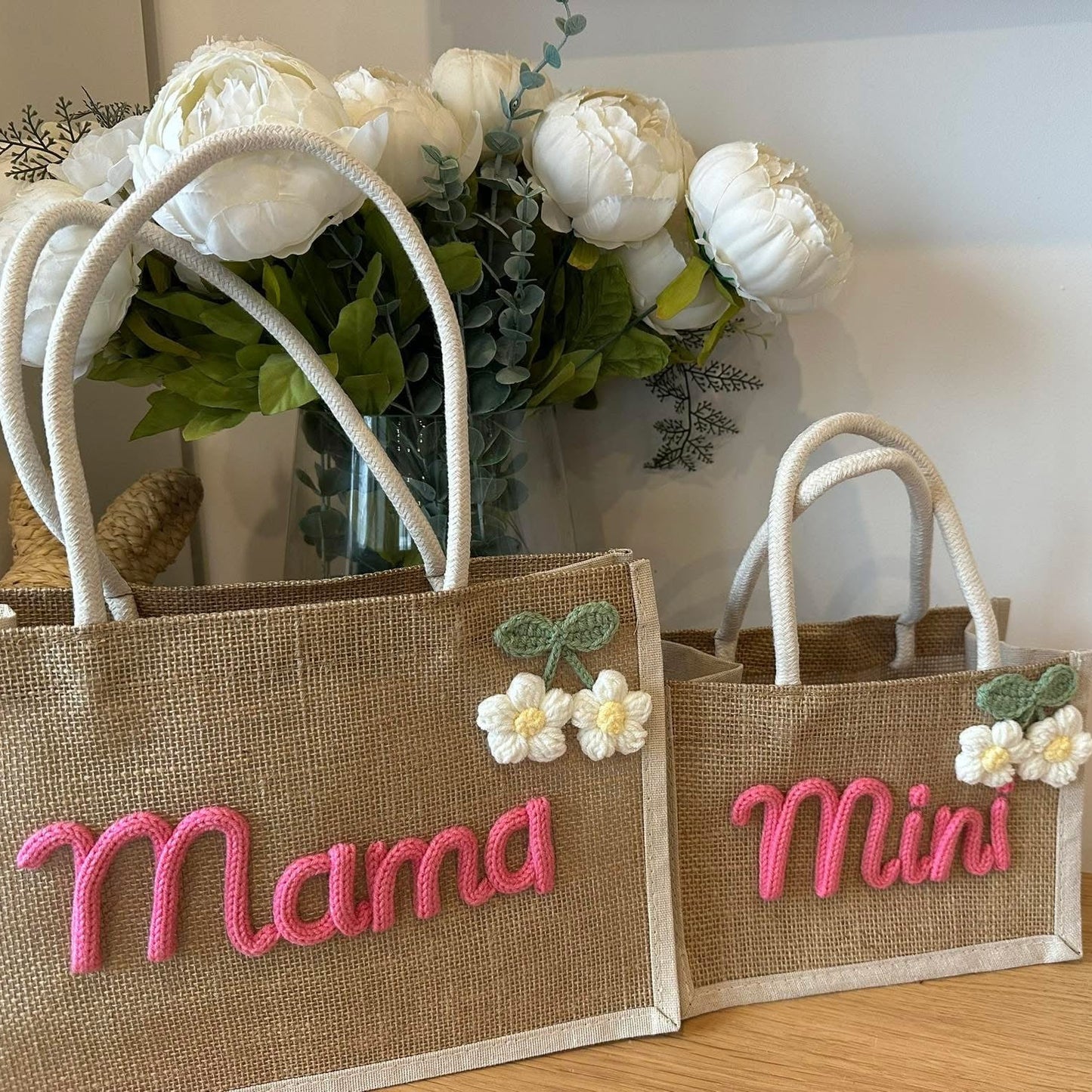 Two jute bags with 'Mama' and 'Mini' text, accompanied by flowers on a wooden surface.