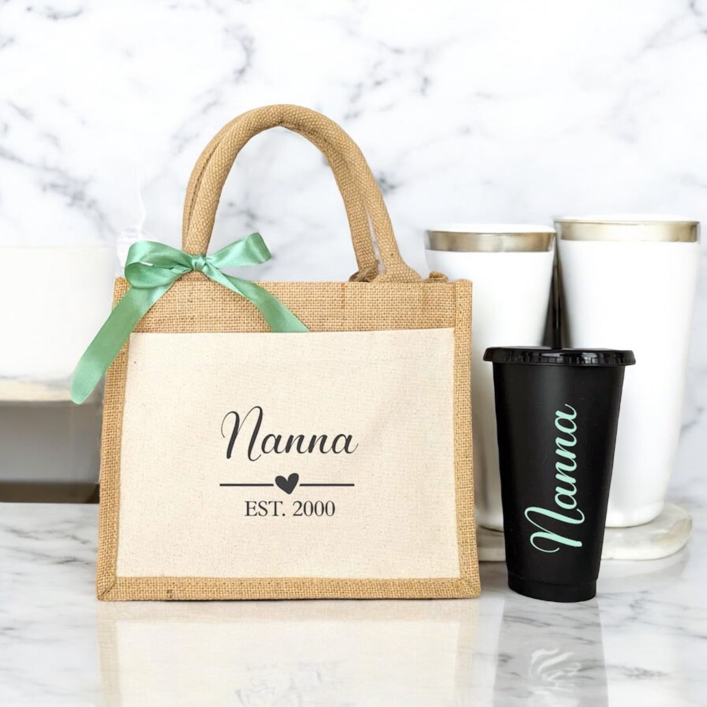 A beige tote bag with the word 'Nanna' printed on the front, alongside a green ribbon and a matching black cup, placed on a marble surface.
