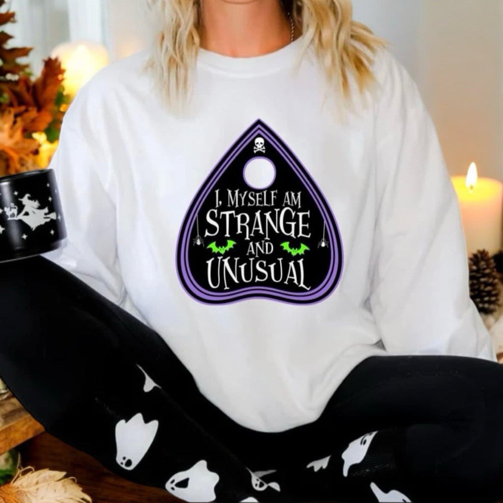 A person sitting and wearing a white long-sleeve sweatshirt with a text 'I must be strange' and a purple graphic on the front, paired with black and white pants.