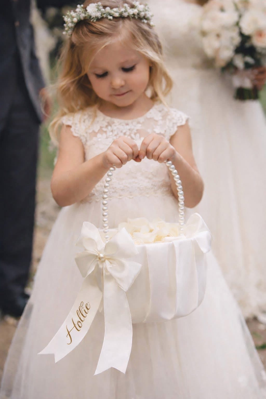 Young girl in a white lace dress holding a basket with 'Holly' on it, standing in front of a blurred background.