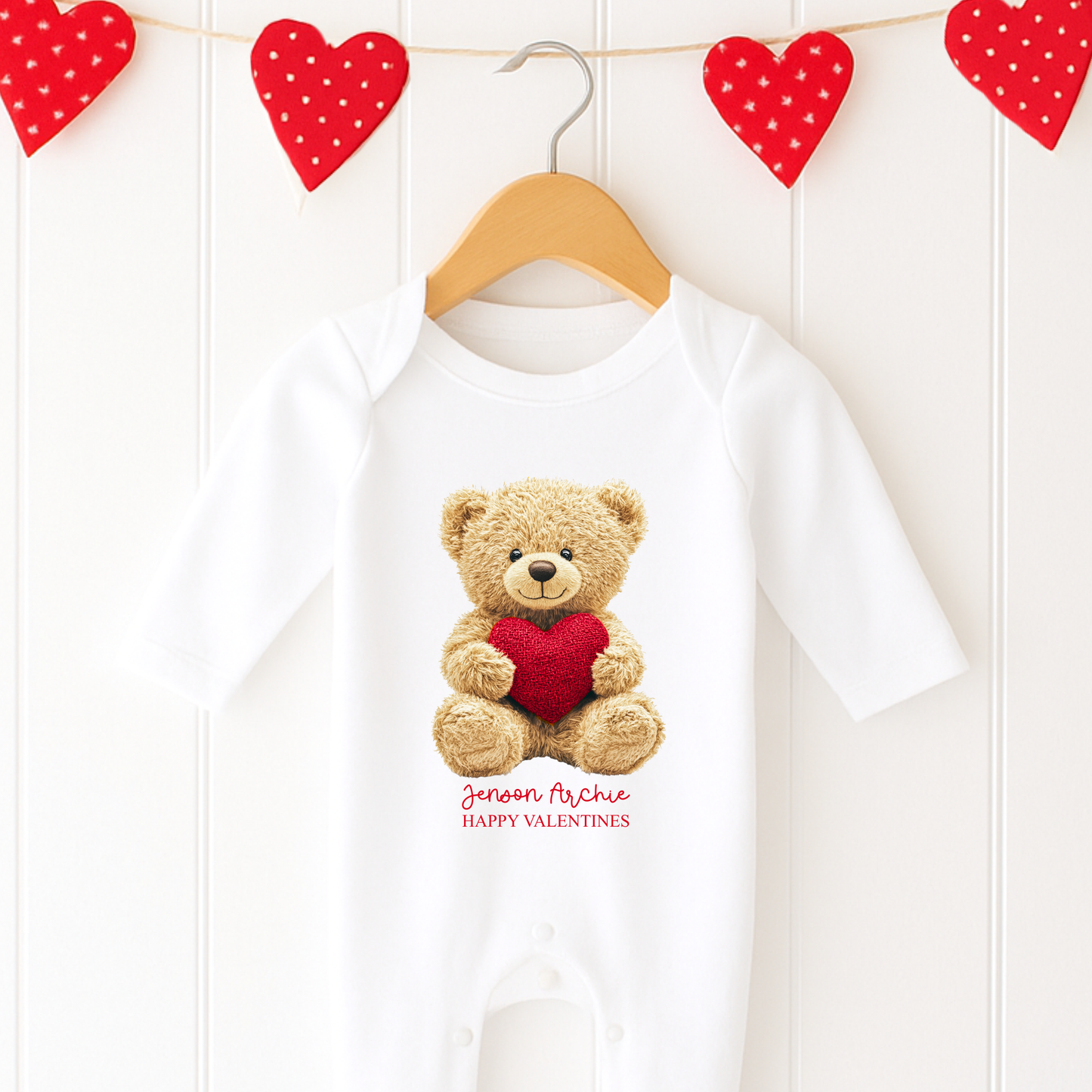 White baby onesie with teddy bear and heart design, hanging on a wooden hanger against a white background with red heart decorations.