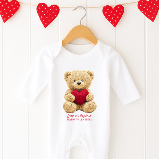 White baby onesie with teddy bear and heart design, hanging on a wooden hanger against a white background with red heart decorations.