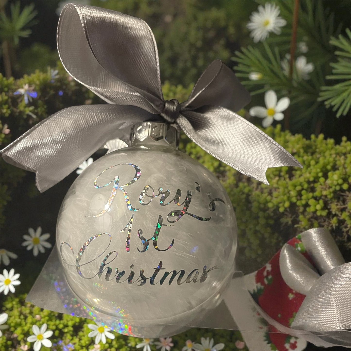 Clear Christmas ornament with 'Baby's 1st Christmas' text and silver ribbon, set against a festive background.