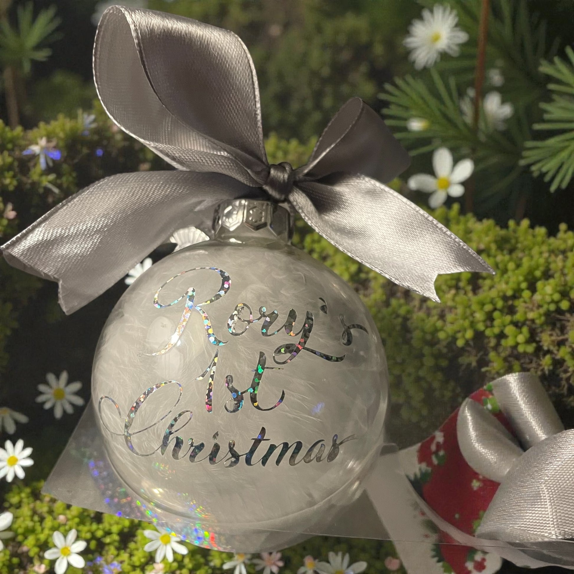 Clear Christmas ornament with 'Baby's 1st Christmas' text and silver ribbon, set against a festive background.