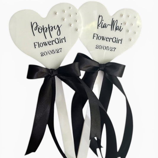 Two heart-shaped flower girl sticks with black ribbons and personalized text on a white background.