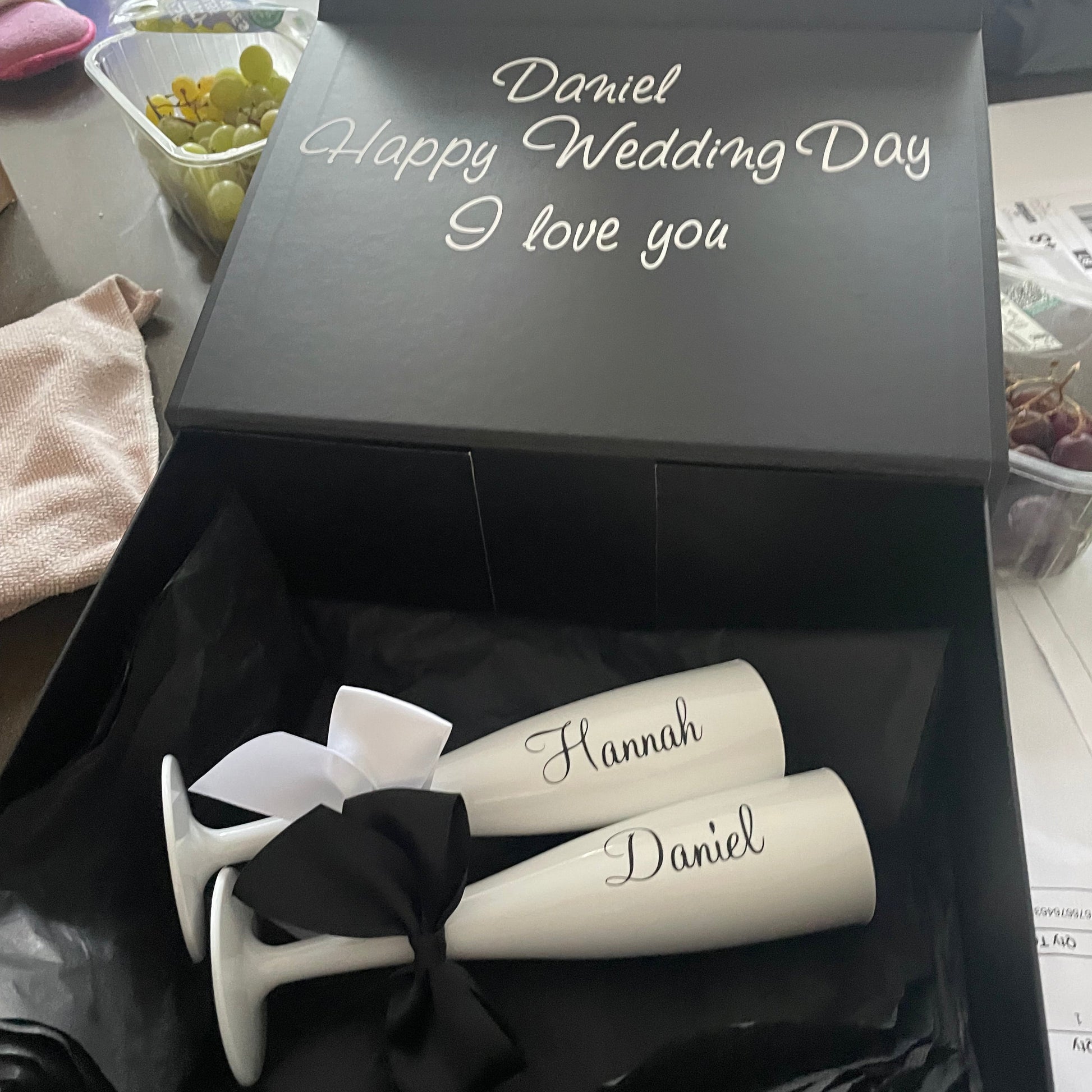 Two white champagne flutes with personalized names in a black box labeled 'Happy Wedding Day'.