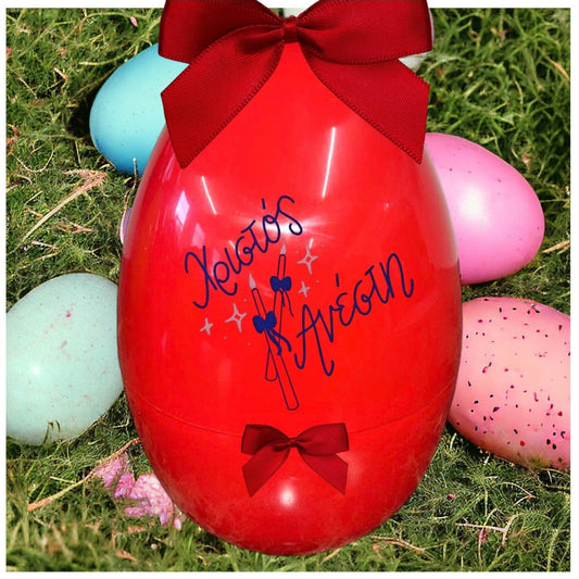 A large red fillable Greek Easter egg with the text 'Happy Easter' written in Greek, accompanied by a red bow and a matching cup, displayed on a grassy surface with other colorful eggs in the background.