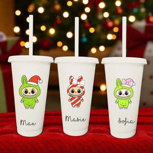 Three white tumblers with cartoon characters and names on a red surface with a blurred Christmas tree in the background.